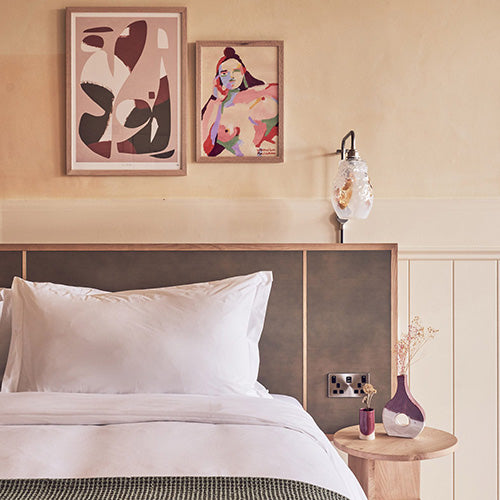 A neatly made bed with white pillows, adjacent to a nightstand, features glass wall pendent above, showcasing simple and elegant interior design.