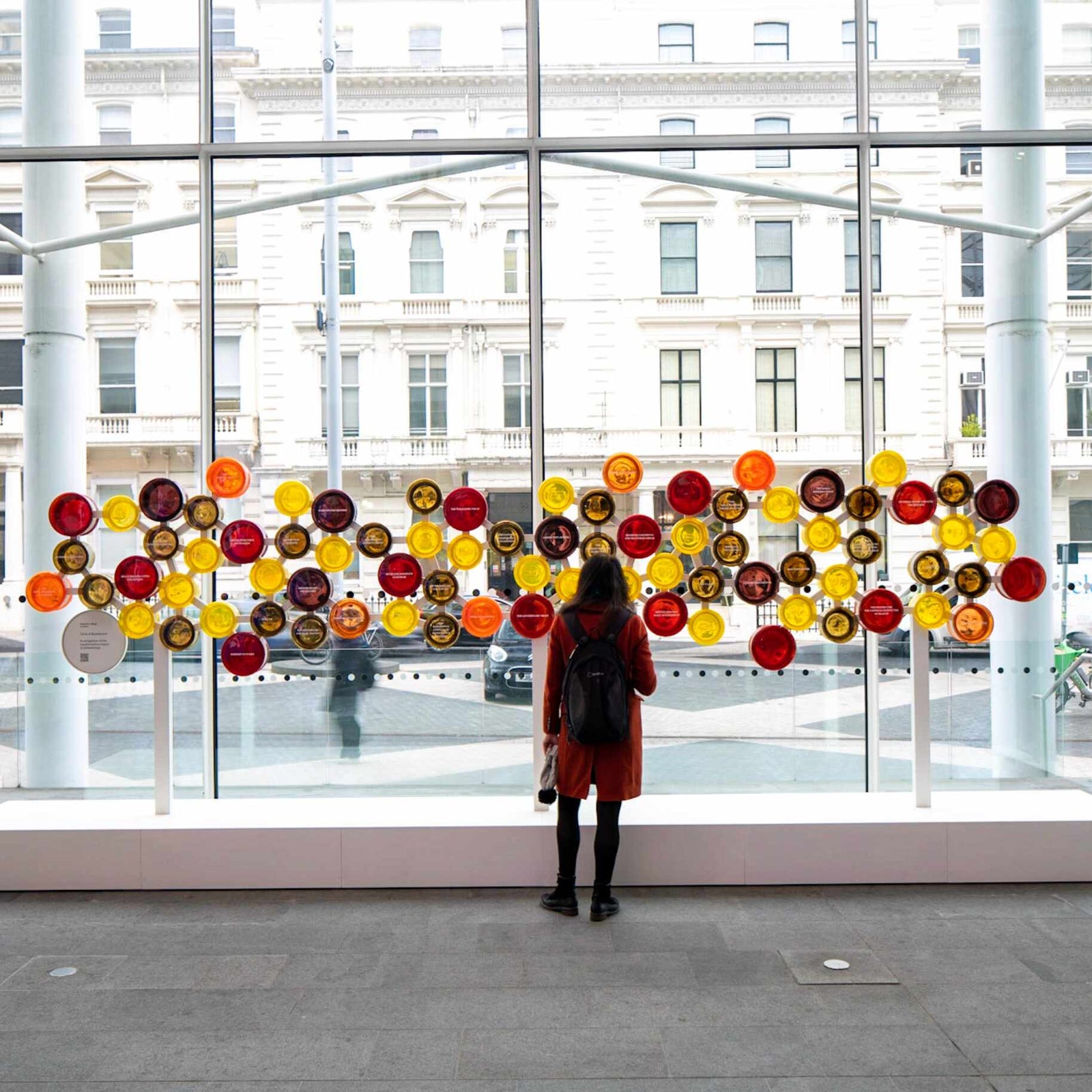 A person in a backpack admires a wall displaying colorful glass plates and circles, standing on a tile floor, in an urban setting.